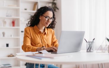 Middle Eastern Lady Using Laptop Working Online Wearing Eyewear Sitting At Workplace In Modern Office. Remote Job, Technology And Career Profession Concept. Side View